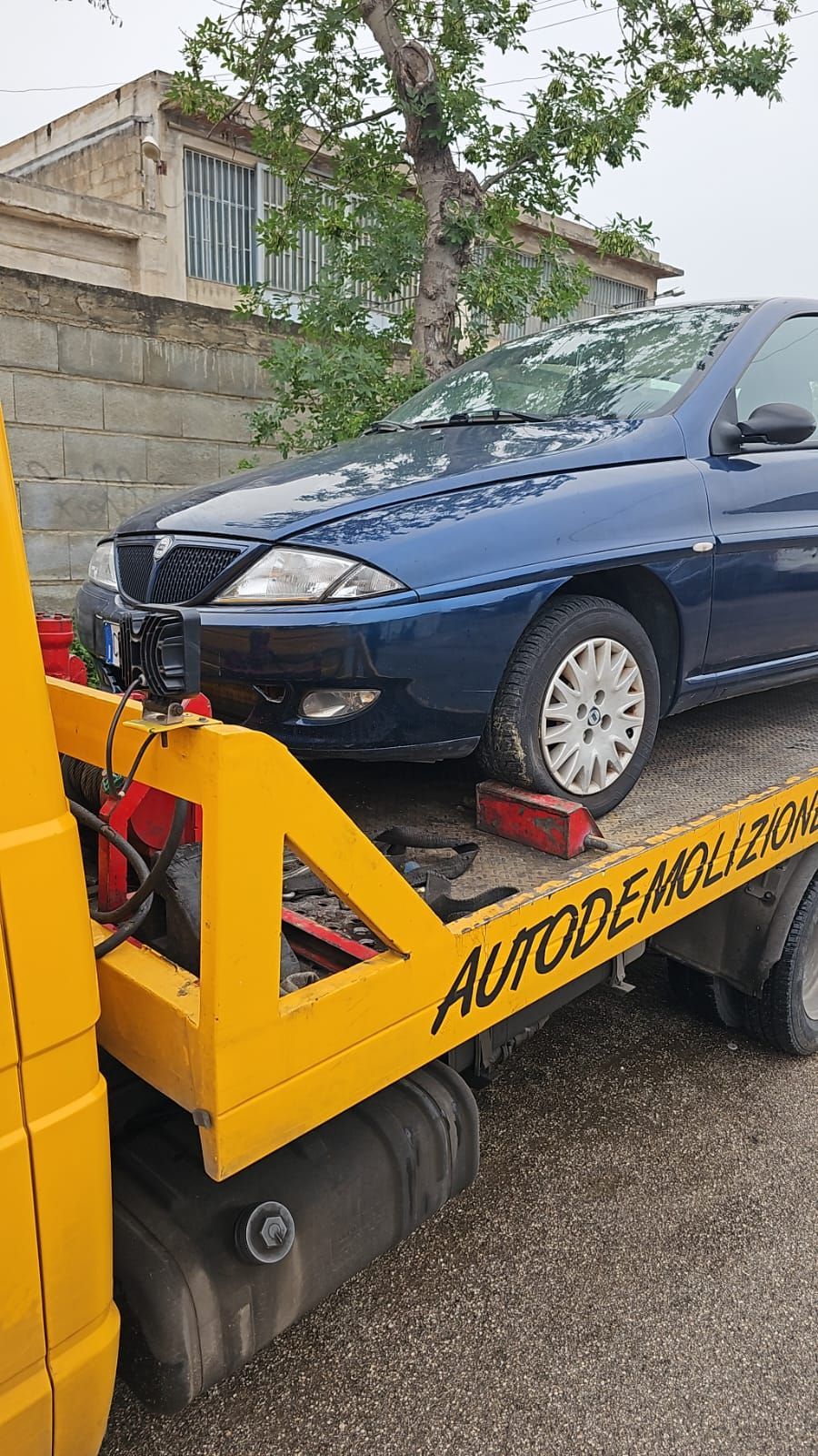 Una Lancia blu è appoggiata sul pianale di un carro attrezzi giallo, fuori da un deposito di rottami.
