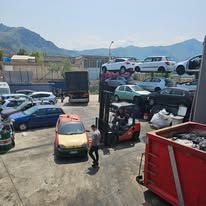 Una persona si trova vicino a un carrello elevatore in un deposito di rottami di auto pieno di vari veicoli, tra cui diverse auto impilate.