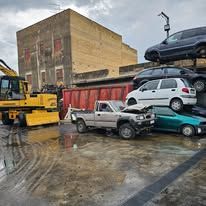 Un escavatore giallo, un pick-up argentato e diverse auto accatastate in un deposito di rottami con un grande edificio sul retro.