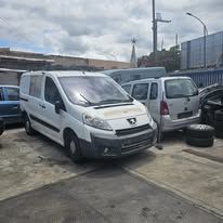 Un furgone Peugeot bianco parcheggiato in un deposito di rottami all'aperto accanto a un minivan argentato, con un'auto blu visibile sullo sfondo.