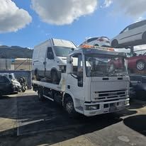 Un carro attrezzi con pianale trasporta un furgone bianco in un deposito di veicoli sinistrati, dove sono parcheggiate altre auto.