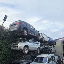 Diverse auto sono accatastate verticalmente in una pila precaria all'esterno di un edificio, sotto un cielo nuvoloso.