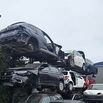 Una pila di auto rottamate in uno sfasciacarrozze, tra cui veicoli scuri, bianchi e rossi, ammucchiati su più livelli.