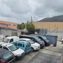 In un parcheggio all'aperto, accanto a una recinzione di lamiera ondulata blu, si trova una fila di auto e furgoni parcheggiati.