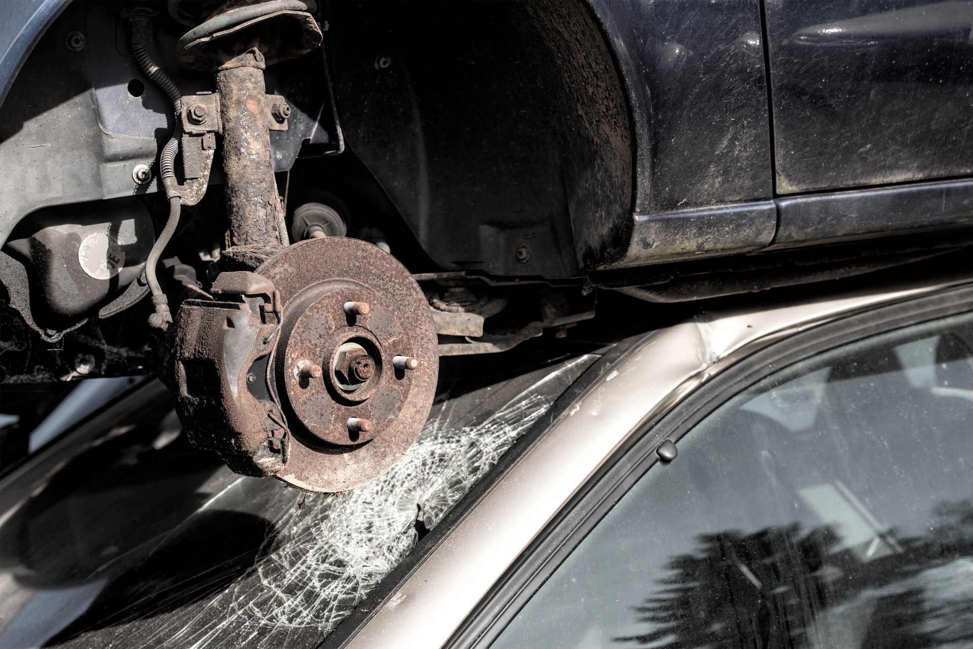 In un deposito di rottami, un gruppo freno arrugginito è sospeso sopra un veicolo argentato con il parabrezza in frantumi.