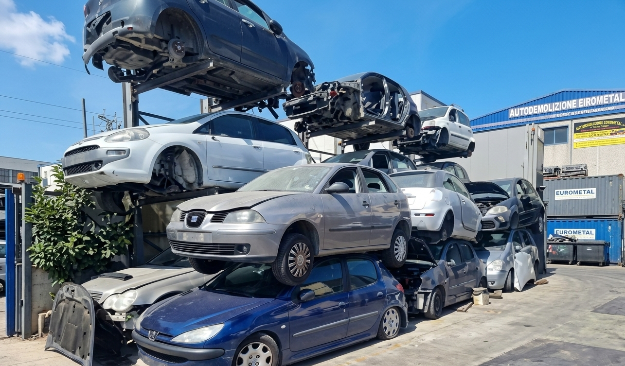 Auto incidentate accatastate in un deposito di rottami, con varie marche e modelli, in un ambiente industriale all'aperto.