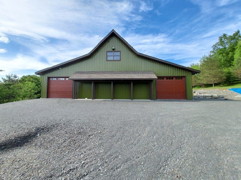 Green wooden building with two red garage doors, under a blue sky, situated on a gravel driveway.