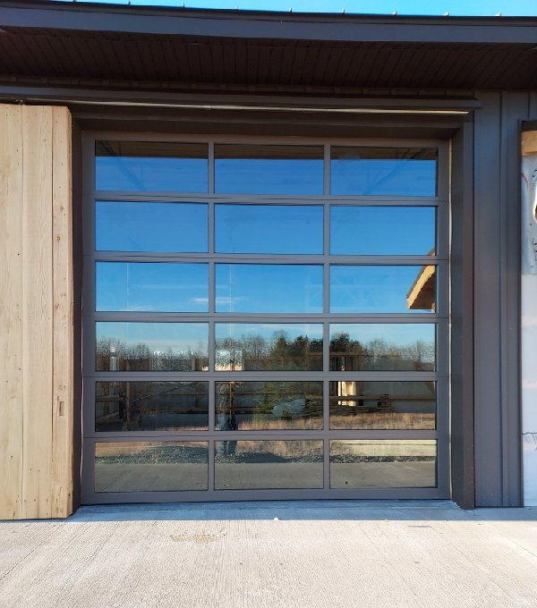 Glass garage door reflecting a blue sky and trees. Grey frame, brown wood siding to the left, and concrete floor.