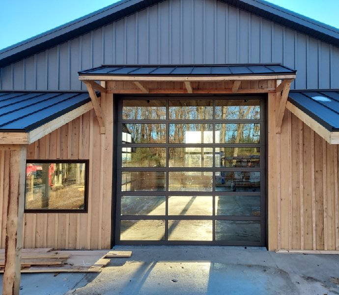 A glass garage door on a rustic building with wood siding. The door has a black metal frame and is reflecting the surrounding trees.