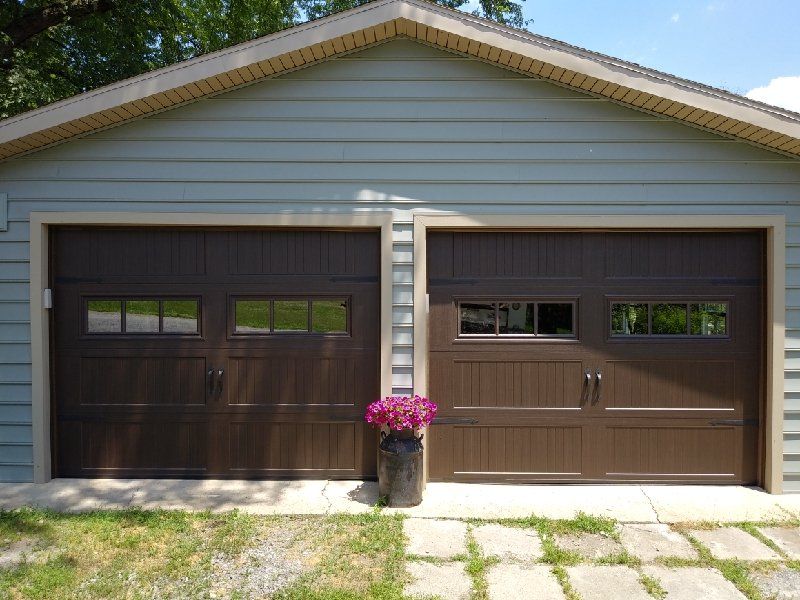Two brown garage doors on a light blue building, with a pot of pink flowers in front.
