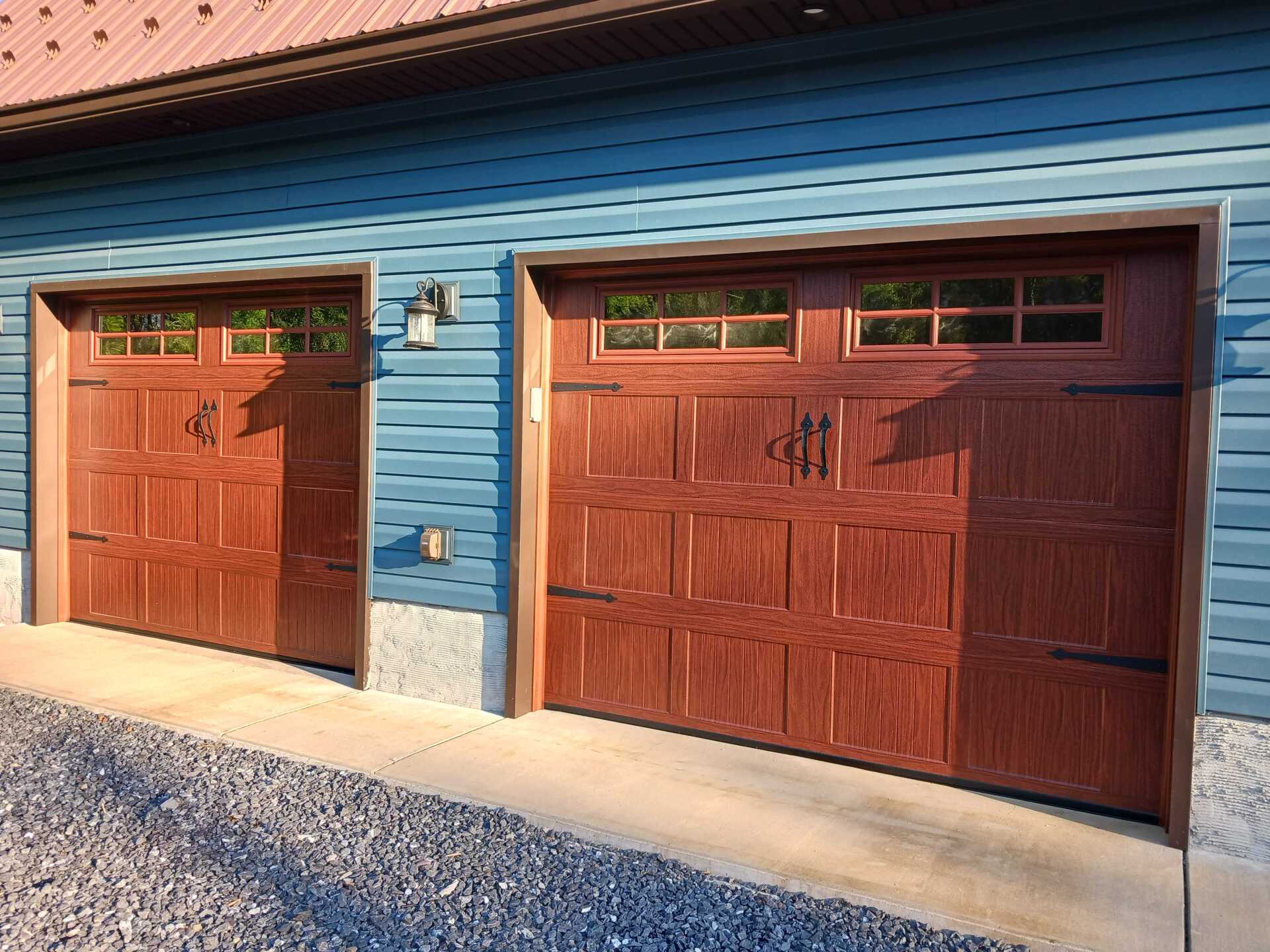 Two matching brown garage doors on a building with blue siding. Each door has decorative iron hardware and a window.