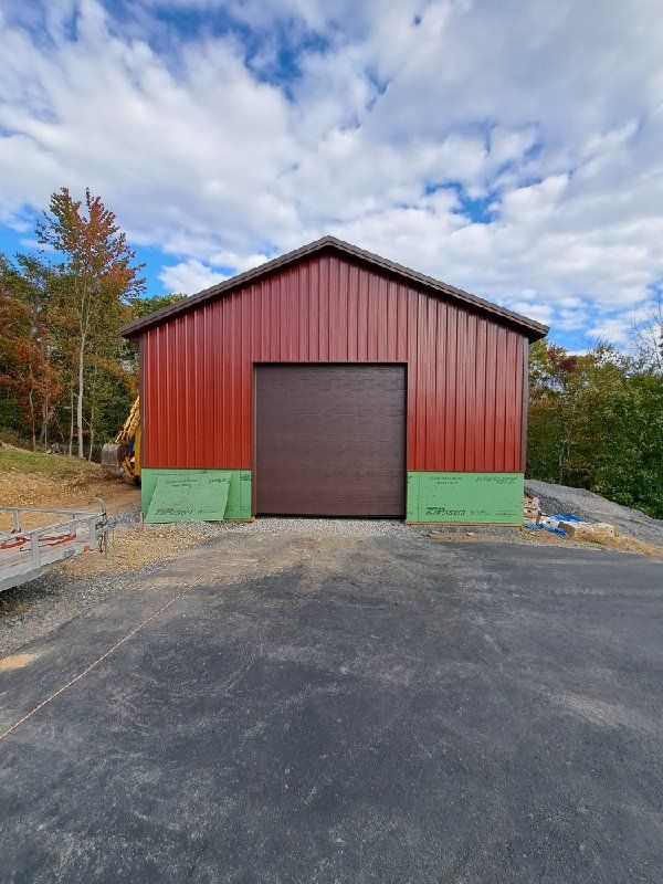 Red metal shed with a brown garage door, green base trim, set on a black asphalt surface under a cloudy blue sky.