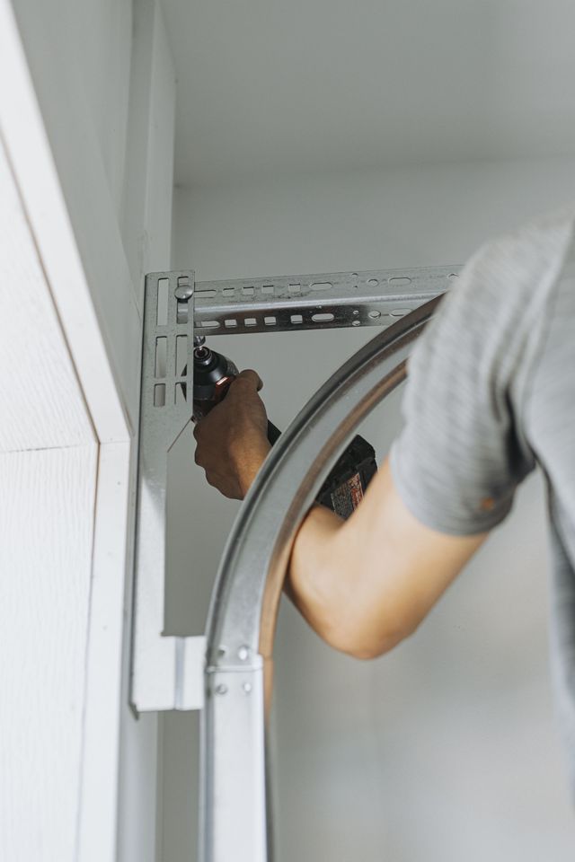 A person in a gray shirt is installing a curved garage door track using a power drill, indoors.