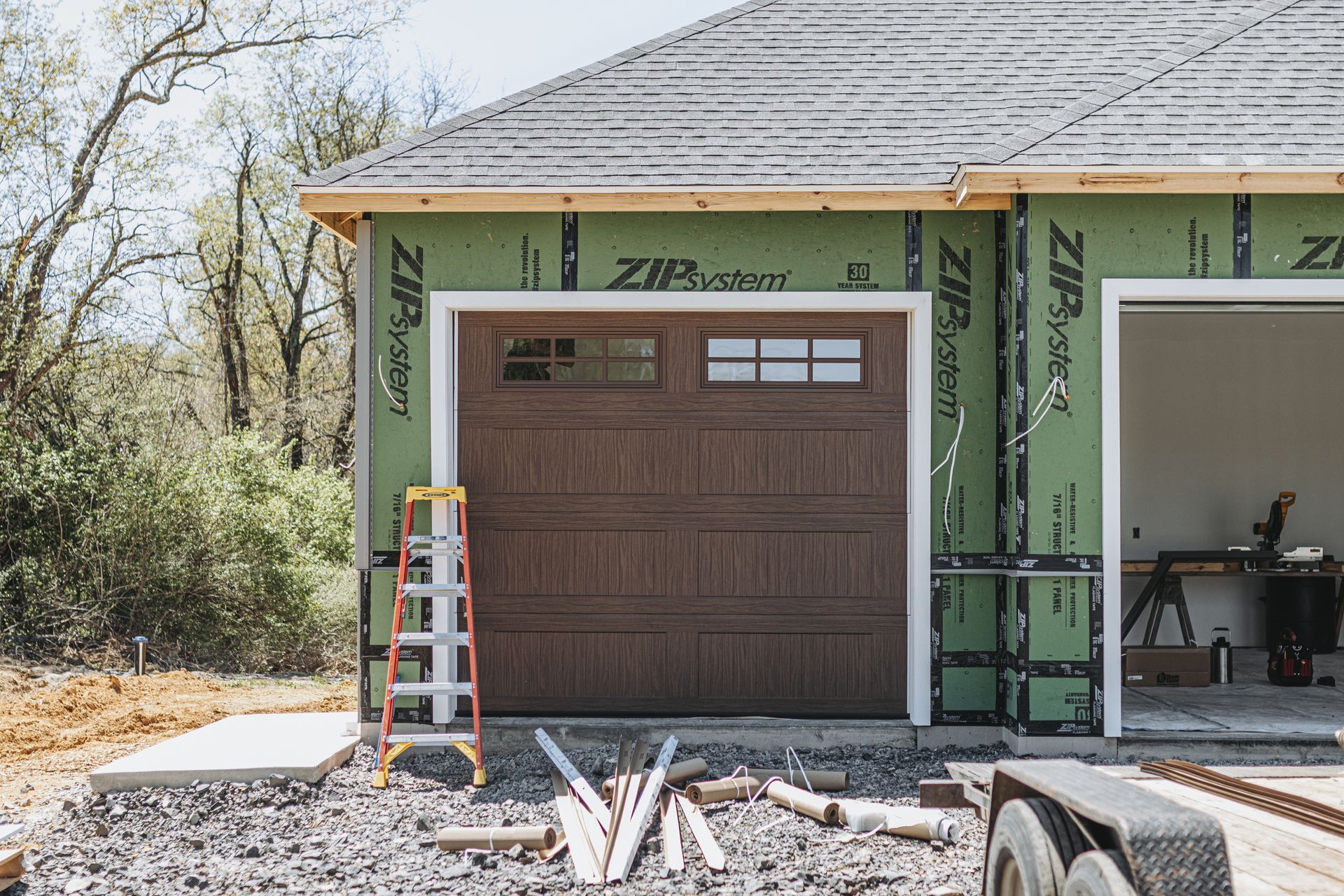 A brown garage door on a partially constructed building with green siding, a ladder, and construction materials in a gravel area.