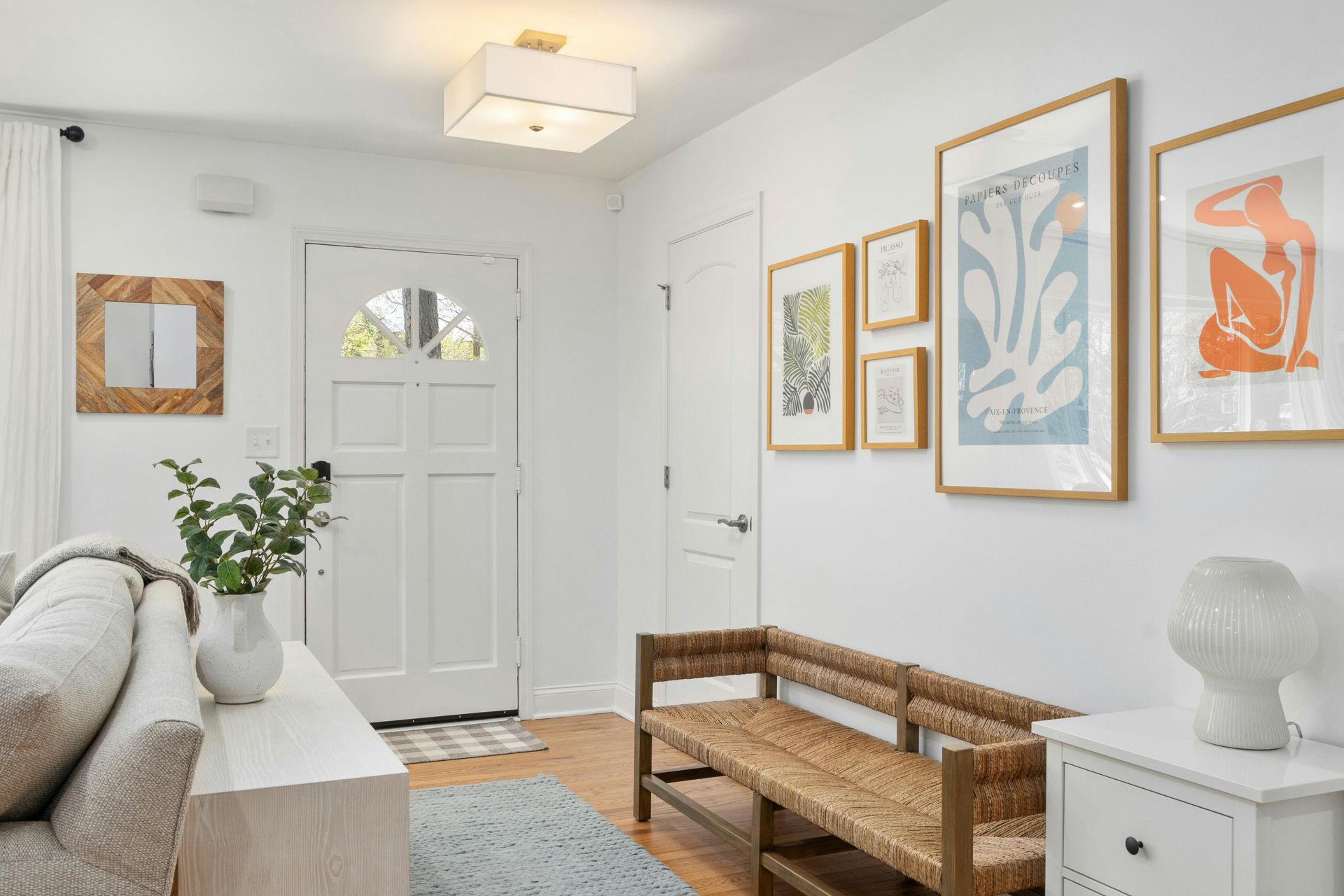 Bright entryway with white walls, a bench, framed art, and a white door. A light blue rug and a sofa are also in the room.