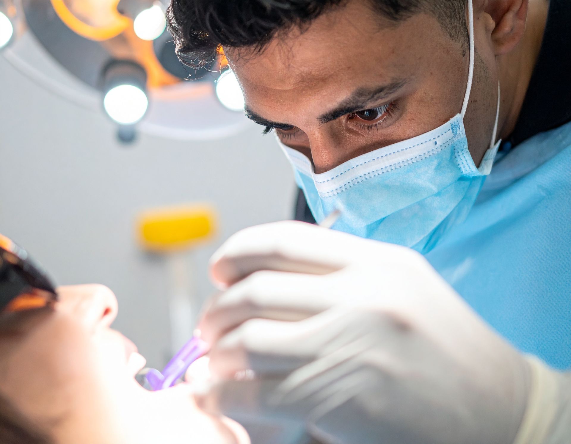 Dentist in blue mask and gloves examining a patient's mouth with a dental light nearby