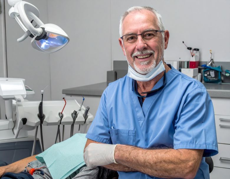 Dentist in blue scrubs, arms crossed, smiling in dental office.