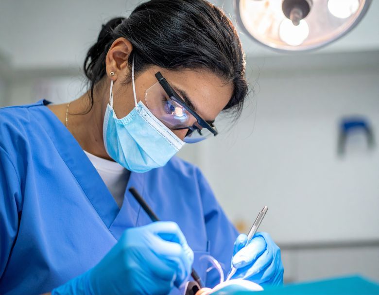 Dentist in blue scrubs, mask, and glasses working on a patient under bright lights in a dental office.