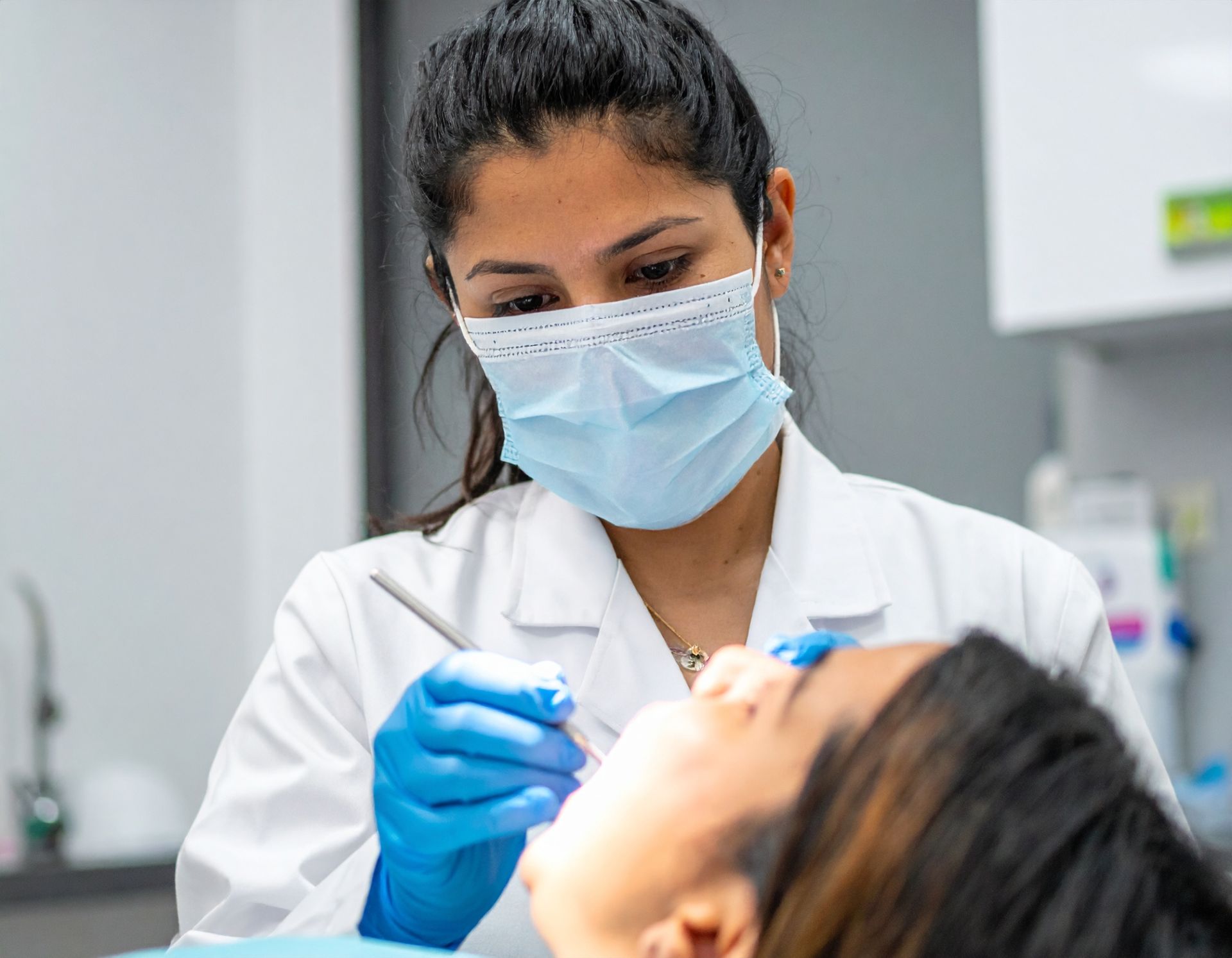 Dentist examining a patient's teeth; both wear masks and gloves. Dentist's white coat and clinic setting.