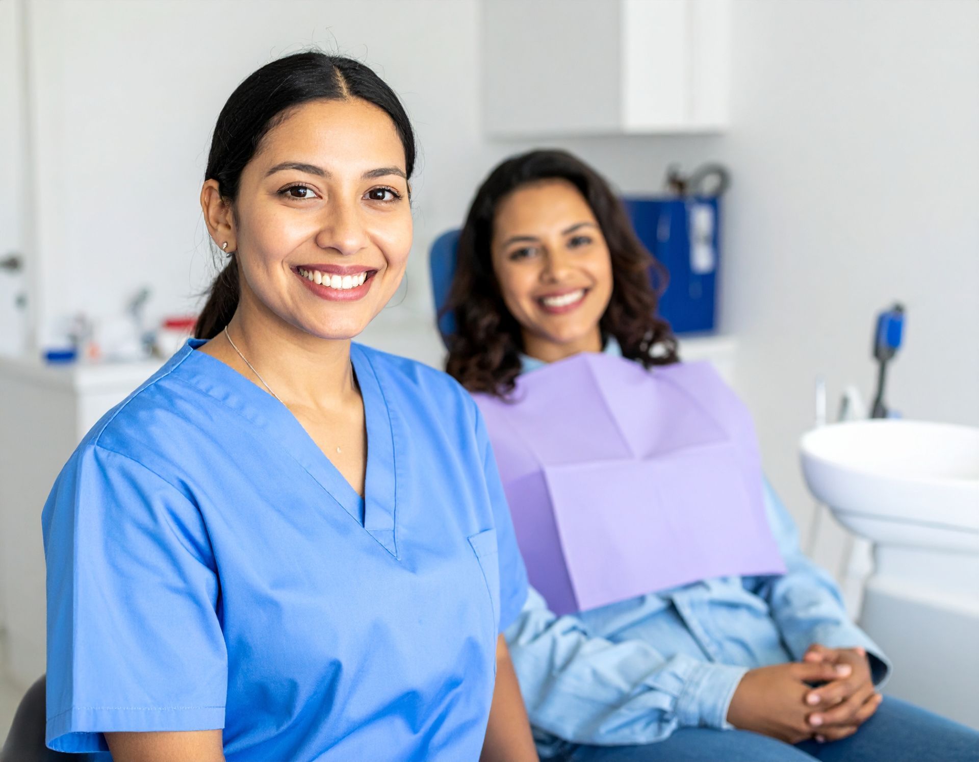 Dental hygienist and patient smiling in a bright clinic; the hygienist wears blue scrubs.