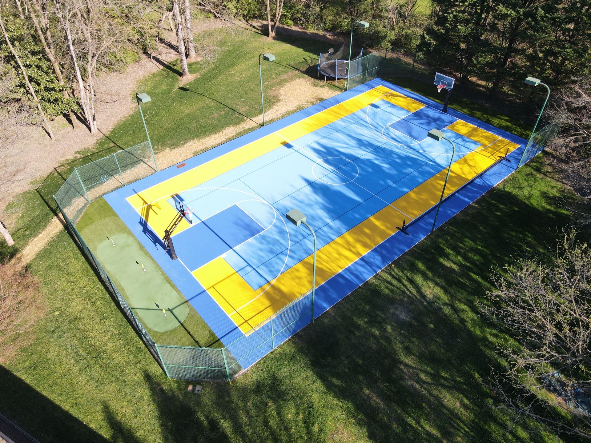 Aerial view of a blue and yellow sports court surrounded by green grass, with a fence and tall trees.