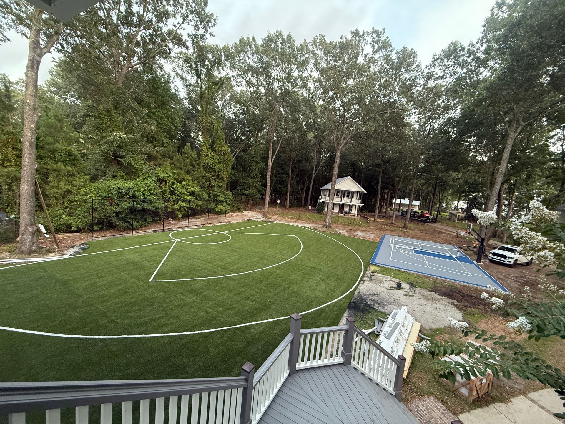 A basketball court on green turf, trees in the background, view from a wooden deck.