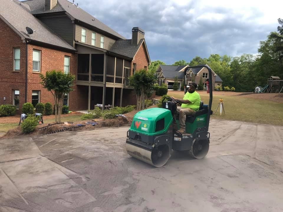 Man operating a green roller on a driveway in front of a house. Cloudy day.