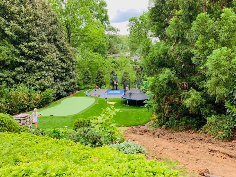 Backyard with putting green, trampoline, and pool surrounded by greenery. Child stands on green.