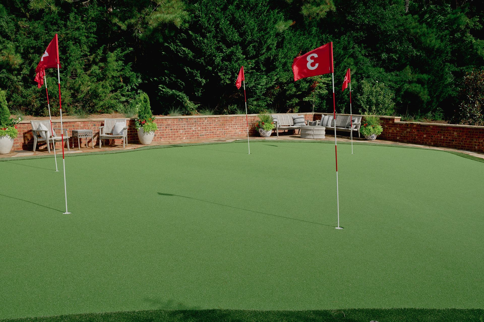 A green putting green with red flags, surrounded by brick wall and greenery.