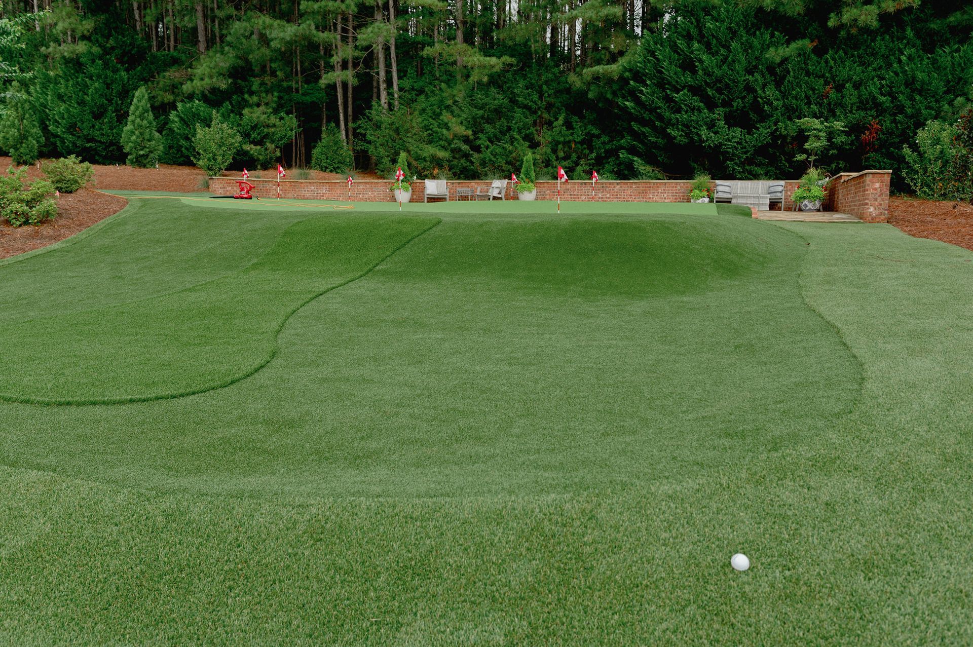 Green putting green with white golf ball, surrounded by brick walls and lush greenery.