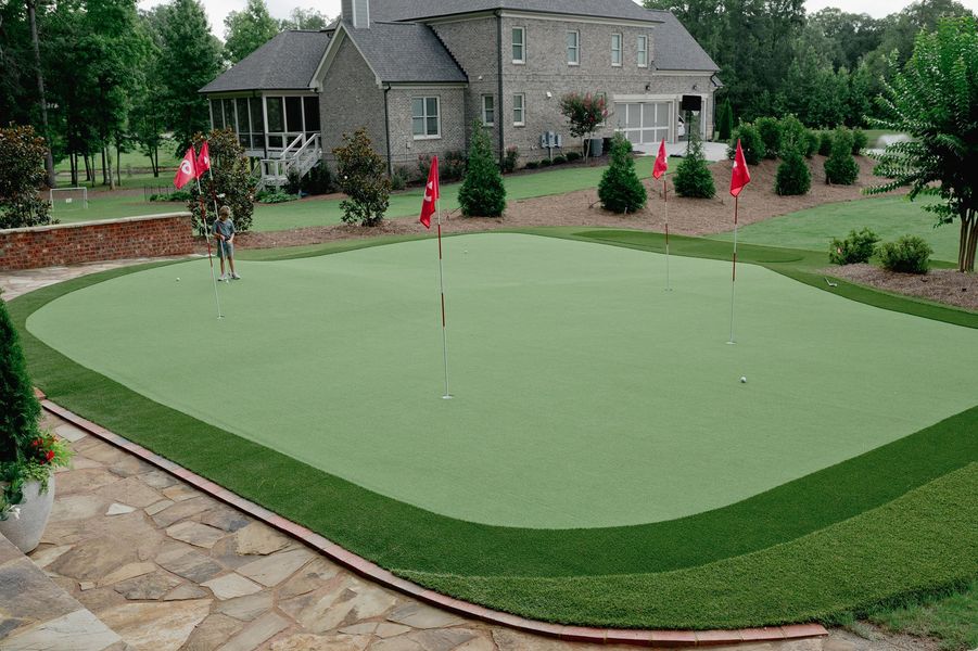 A backyard putting green with flags, surrounded by a dark green border and landscaping, in front of a brick home.
