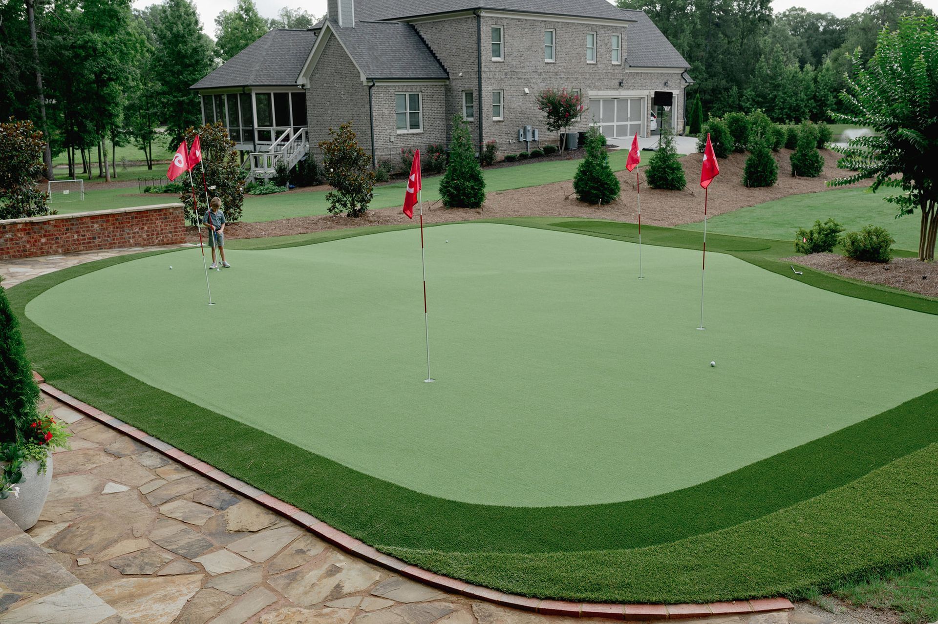 A putting green with red flags and multiple holes in a backyard setting next to a stone patio.