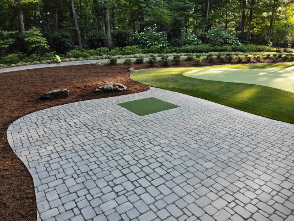 Stone patio curves to lawn and putting green, surrounded by mulch and trees.