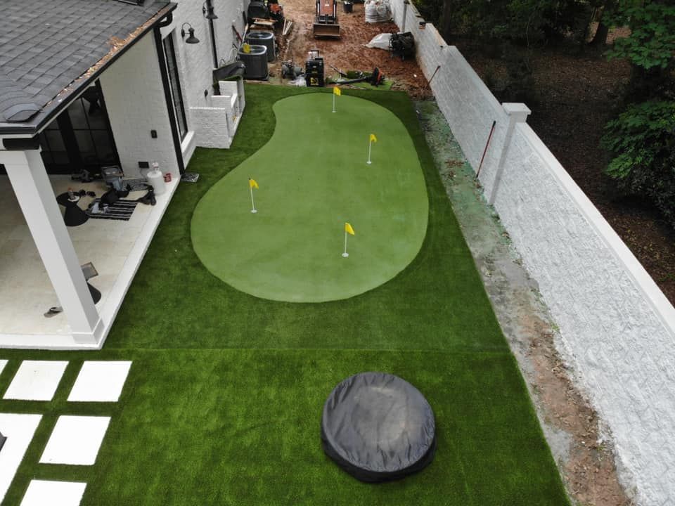 Backyard golf green with several flags, surrounded by green turf and a white fence.