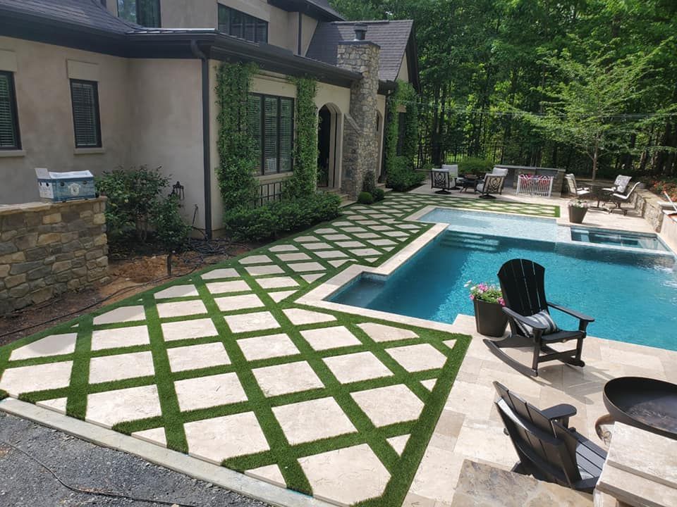Backyard pool area with a grid of grass and stone, surrounded by trees and patio furniture.