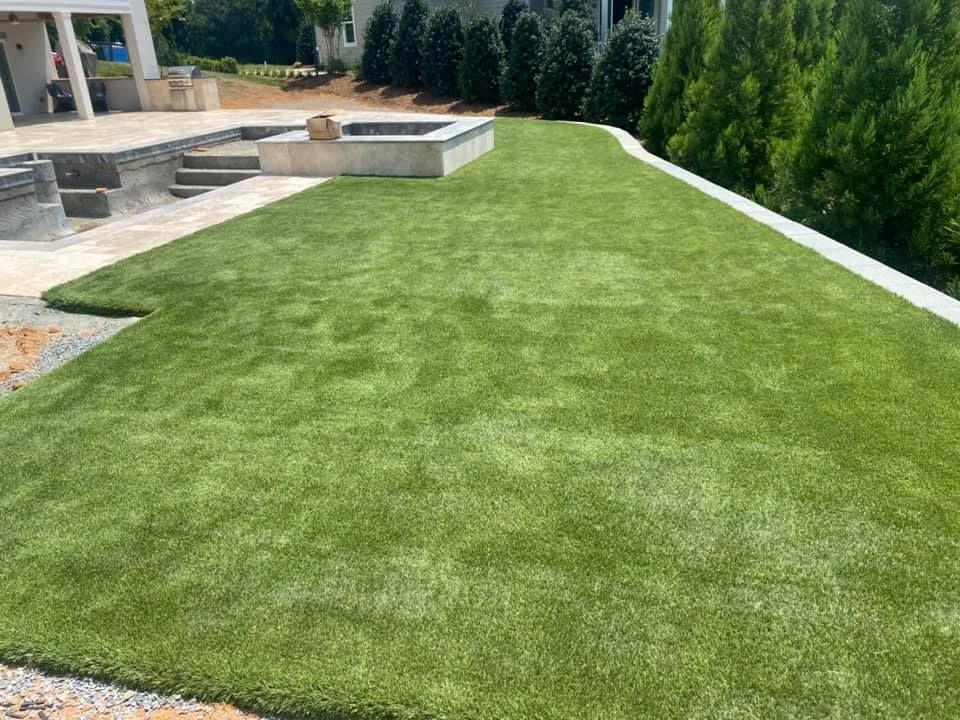 Green artificial turf lawn next to a patio and low wall with evergreen trees in the background.