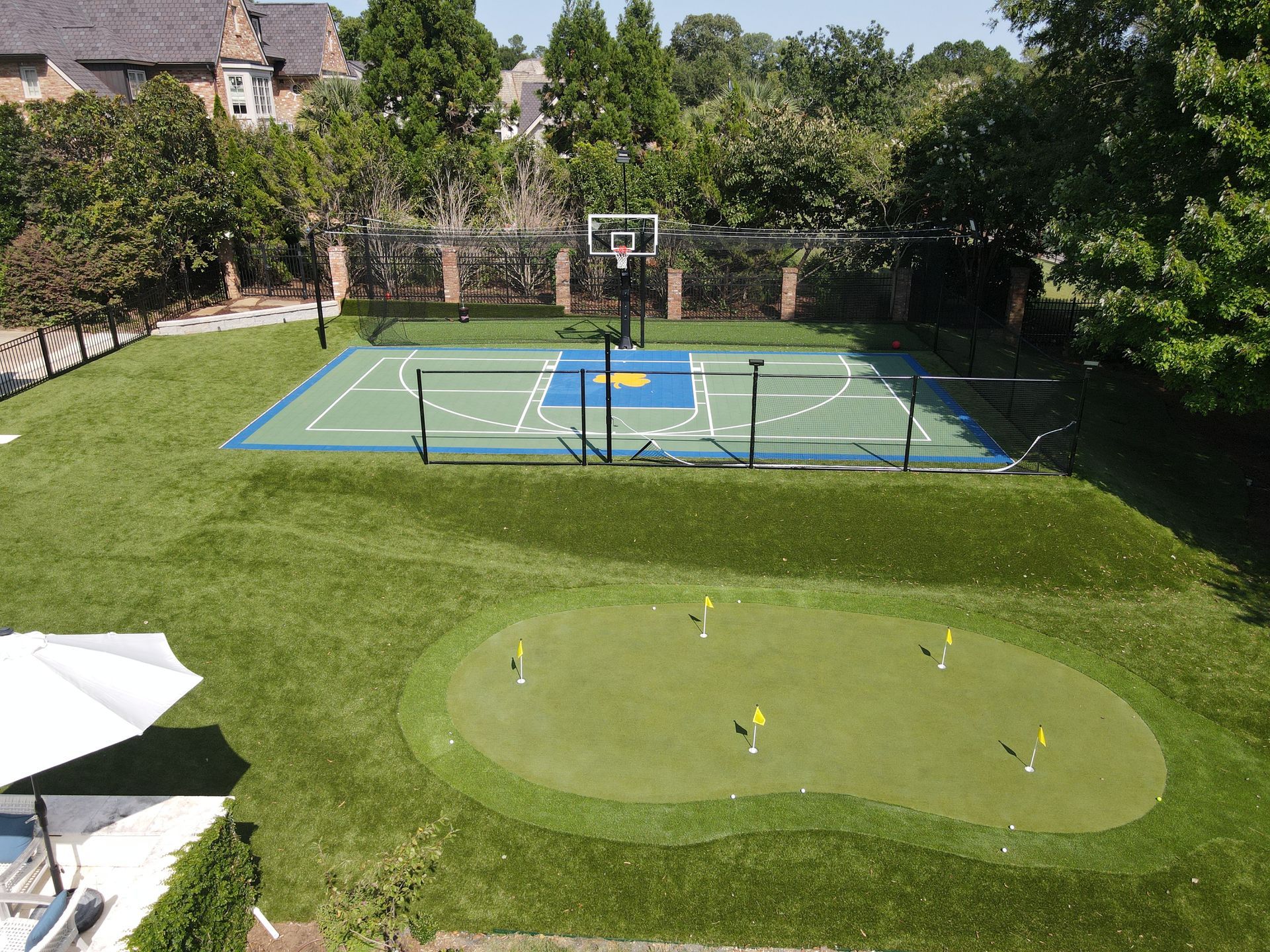 Backyard with basketball court and putting green, surrounded by green grass and trees.