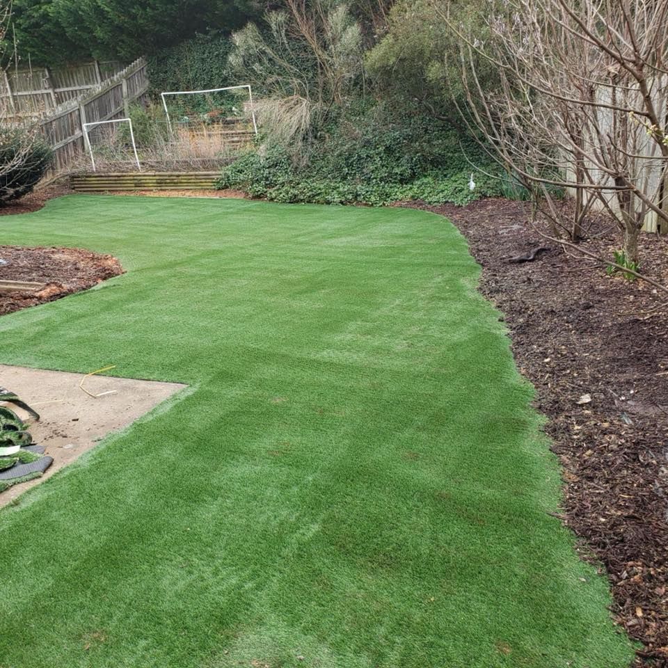 Newly laid green artificial turf in a backyard with a retaining wall and sparse trees.