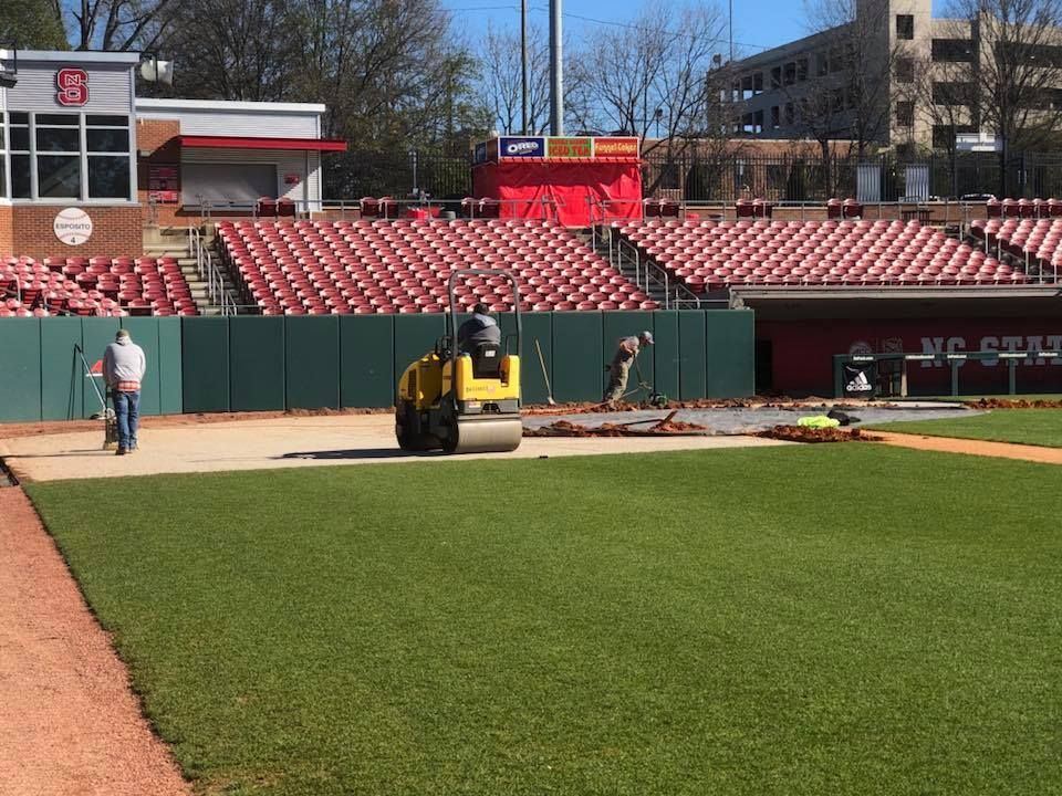 Baseball field maintenance: workers compacting soil and laying grass near red stadium seating on a sunny day.