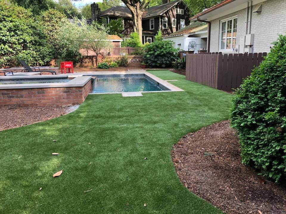 Backyard with a pool and artificial green grass surrounded by landscaping and fencing.
