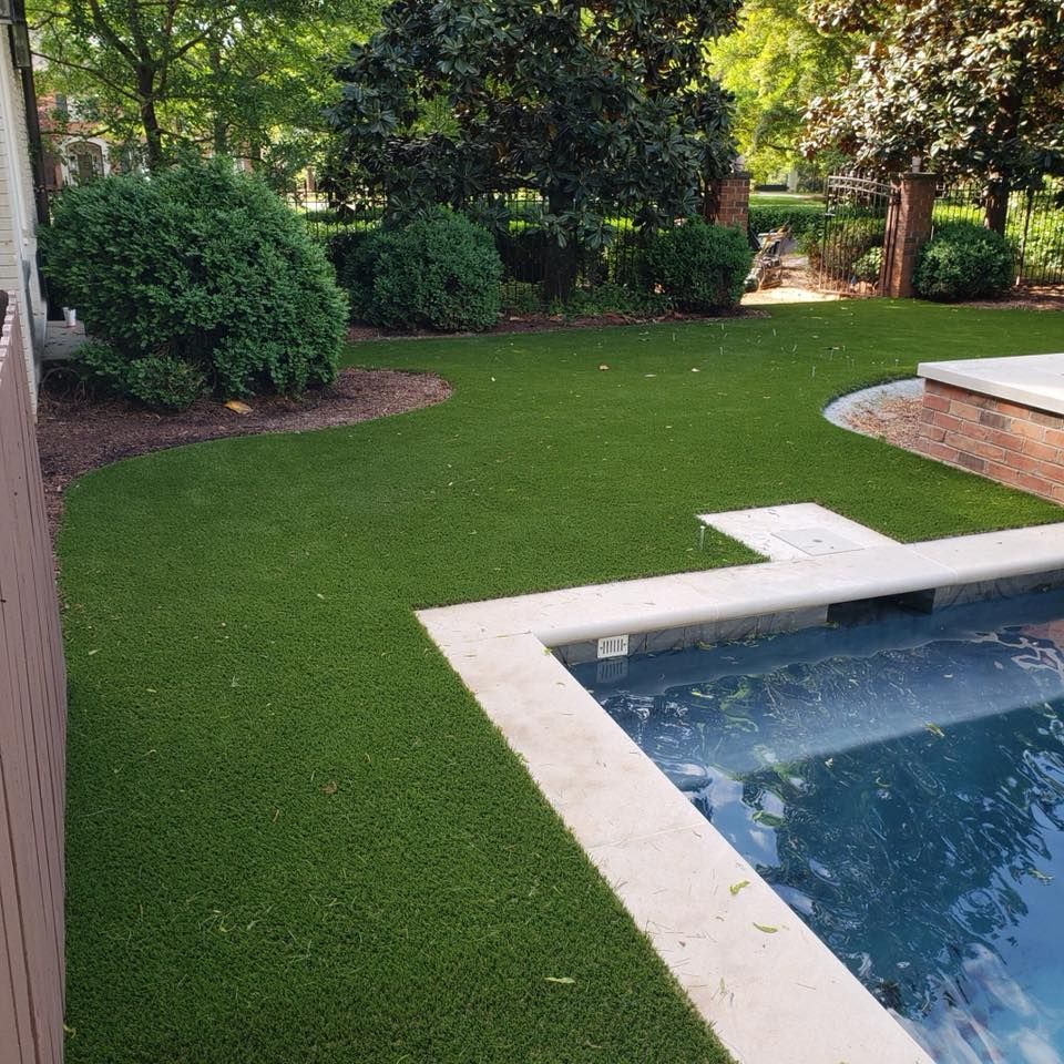Artificial turf surrounds a blue pool in a backyard with bushes and trees.
