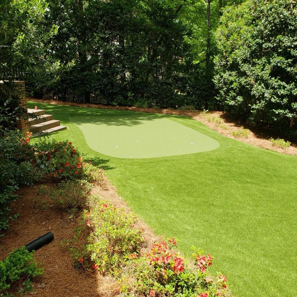 Artificial turf backyard with a putting green, surrounded by trees and bushes.