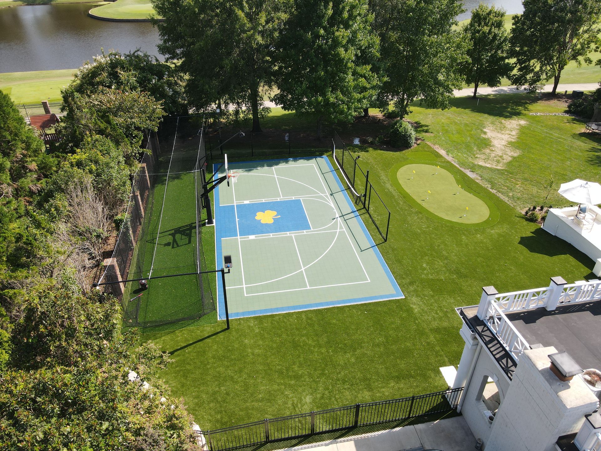 Aerial view of a backyard with a basketball court, putting green, and lake, surrounded by green grass and trees.