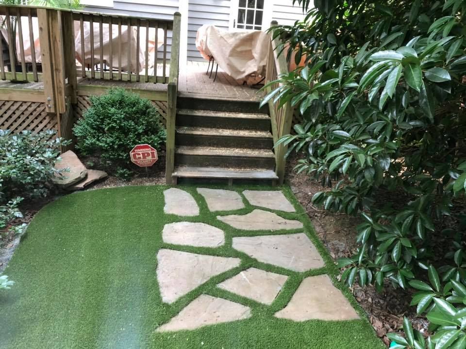 Stone path leads to wooden deck with covered furniture; artificial turf lawn.