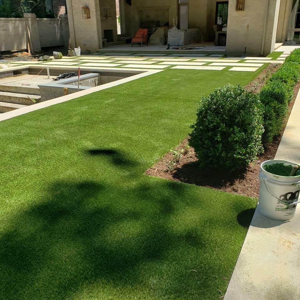 Green artificial turf with shrubs beside a concrete patio and building.