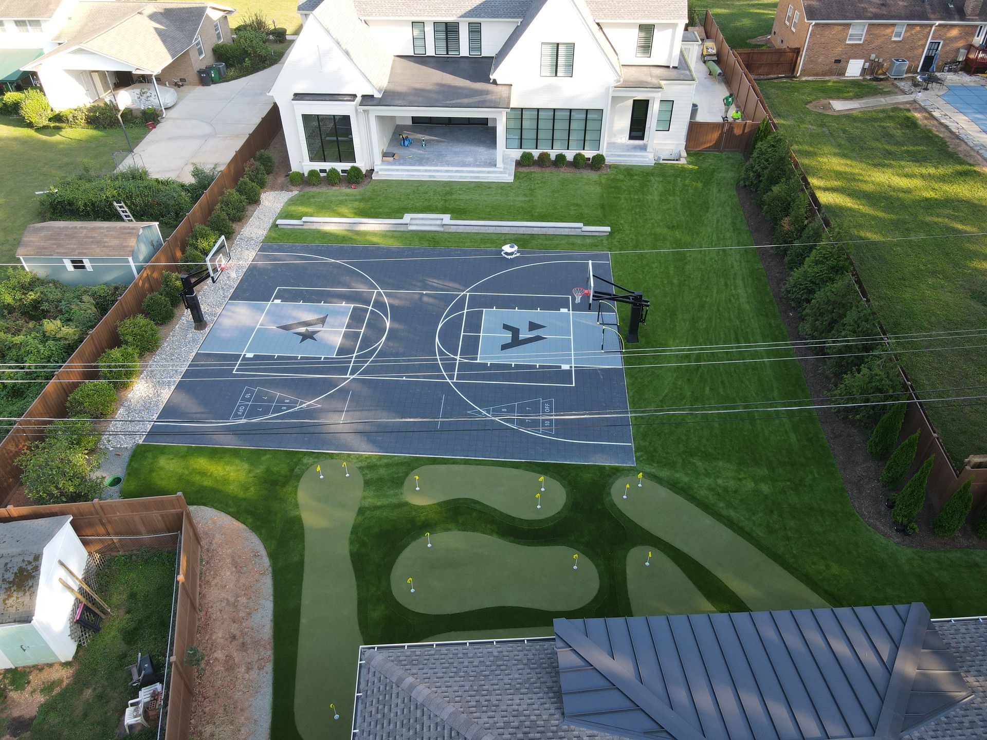 Aerial view: backyard with basketball court, putting green, large white house, green grass.