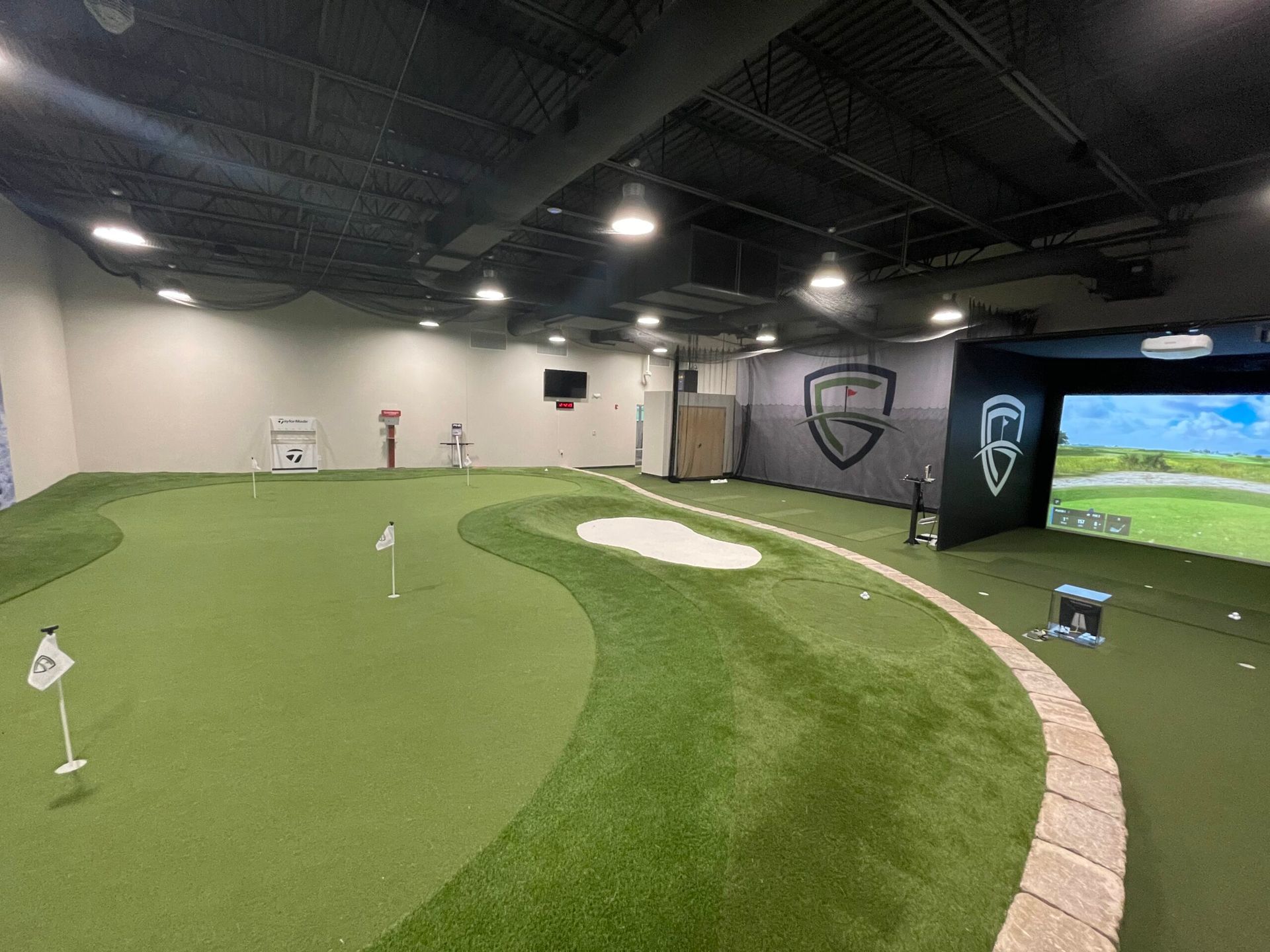 Indoor golf simulator with putting green, sand trap, and screen displaying a golf course.