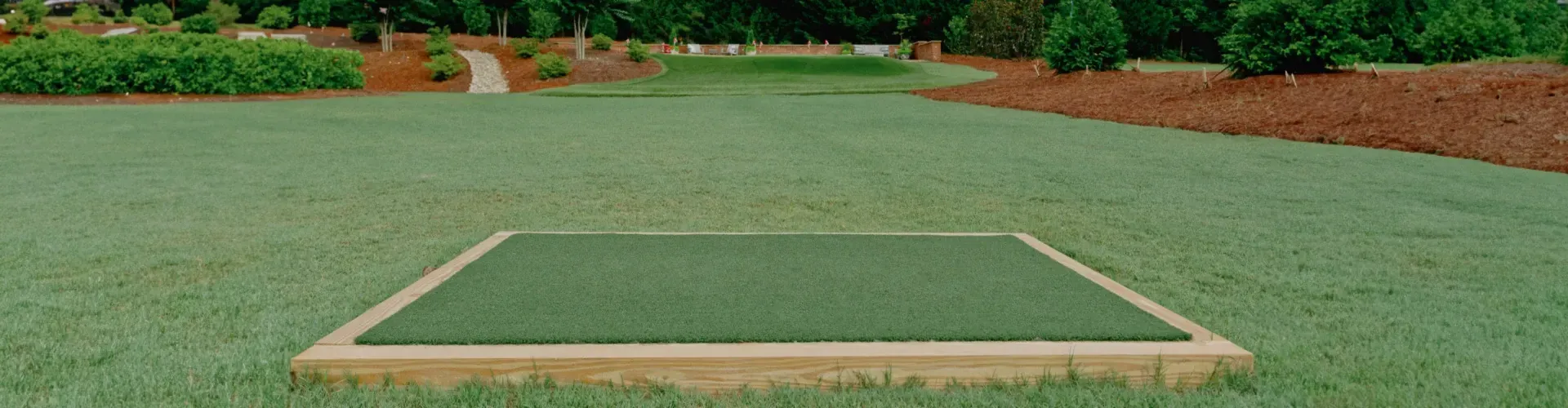 A golf tee box in a grassy field, surrounded by trees and a path.