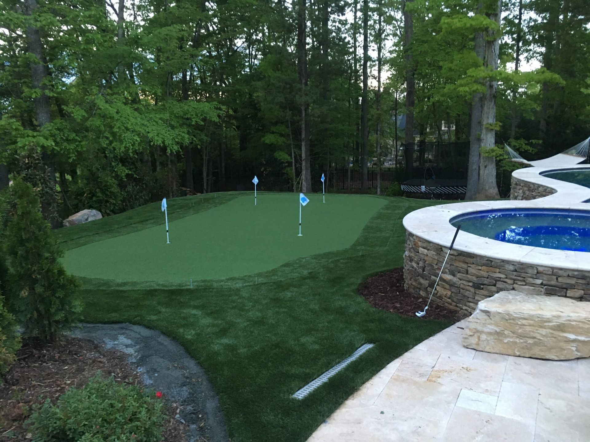 A backyard putting green with blue flags and a pool in a wooded setting.