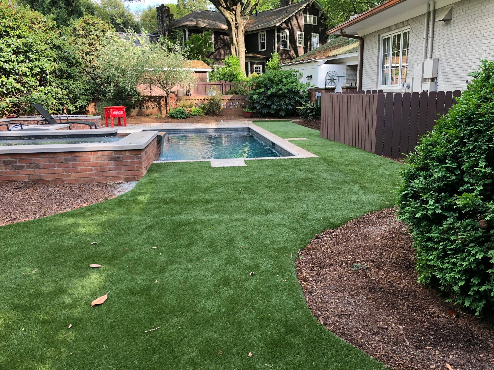 Backyard with a pool, green turf, brick border, brown fence, and surrounding greenery.